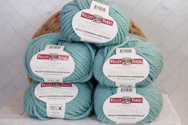 A tower of five skeins of Valley Yarns Superwash Super Bulky in a light teal color. It sits on a flat white blanketed surface propped up as a product photo against a white tile background.