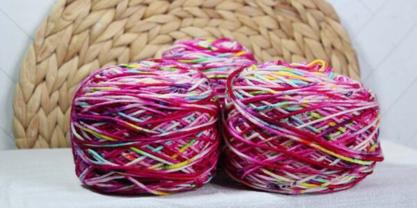 Three cakes of yarn wound and laying on a display table. Woven brown circle behind the yarn, and the table has a white cloth over it. Yarn is variegated rainbow colors, but predominant pink.