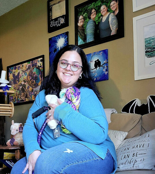 Christina sitting on an armless couch holding three hanks of yarn and her cane tucked between her legs, smiling at the camera in a light blue sweatshirt and jeans with glasses and hoop earrings.